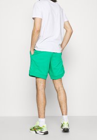 Tyrkysové kraťasy s bočními kapsami, elastickým pasem a viditelnými stehy, spárované s vzorovanými sportovními botami a bílými ponožkami.