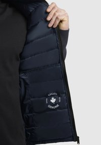 Gilet matelassé marine, avec un design quilté et une ouverture zippée. L'intérieur présente un écusson logo avec un motif de feuille et un texte associé.