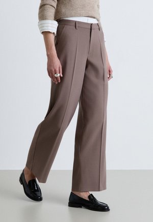 Personne portant un pantalon large taupe, des mocassins noirs, un pull beige avec des manches de chemise blanches rayées visibles, et des bagues argentées aux deux mains.