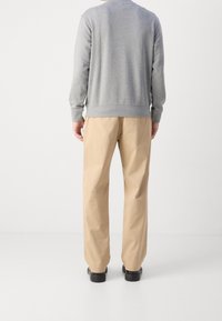Persona vistiendo pantalones beiges, zapatos negros y una camiseta de manga larga gris, de pie de espaldas frente a un fondo blanco liso.