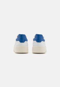 Baskets en cuir blanc avec des accents bleus, présentant une forme arrondie, une pièce arrière texturée et une semelle en caoutchouc crème texturée. Design minimaliste.