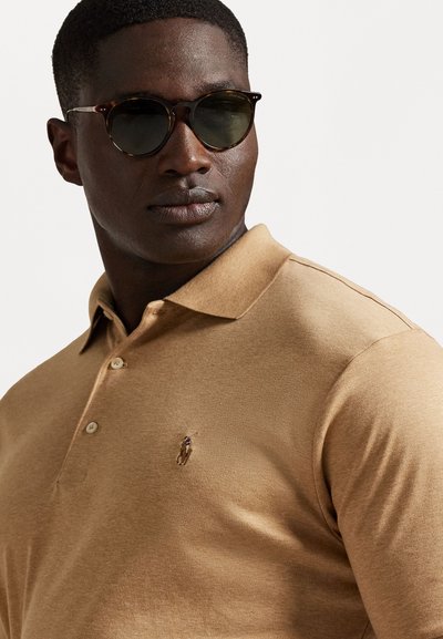 Μπεζ πουκάμισο πόλο με γιακά και τρία κουμπιά. Μικρό λογότυπο στο στήθος. Άτομο που φορά σκούρα, στρογγυλά γυαλιά ηλίου με φιμέ φακούς.