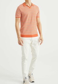 Orange och vit randig kortärmad polo med V-ringning och stickad textur, i kombination med vita byxor och neutrala sneakers.