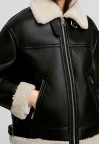 Person i svart läderjacka med vit fårskinnskrag och manschetter, hand i dragkedjefickan, frontdragkedja och spändetaljer synliga.