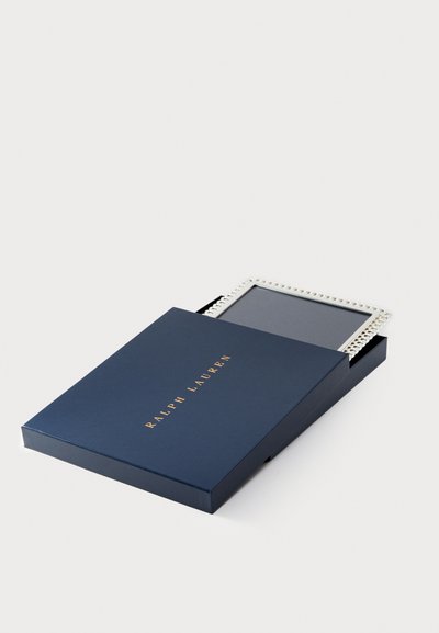 Tamsiai mėlyna dėžutė su auksiniais užrašais, kurioje yra stačiakampis nuotraukų rėmas su dekoratyviu sidabriniu kraštu. Rėmas iš dalies matomas iš atidarytos dėžutės.