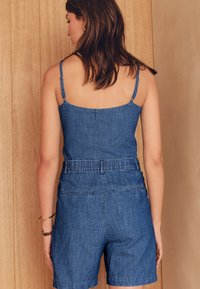 Džinsinis kombinezonas su plonomis petnešėlėmis, suvarstytu liemeniu ir kišenėmis nugaroje. Audinys tamsiai mėlynas, lygios tekstūros, su minimaliais siuvimo akcentais.