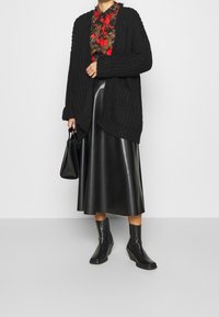 Cardigan negro de punto con apertura frontal, blusa floral, falda de cuero negra de cintura alta y botines negros, sosteniendo un bolso negro.