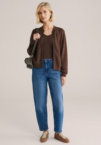 Barna vadászdzseki barna felsővel, kék magasderekú farmerrel párosítva. A barna cipők világos fűzőkkel és texturált mintával rendelkeznek.