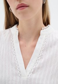 Blusa bianca in tessuto testurizzato, con scollatura a V e rifinitura in pizzo. Il colletto ha un dettaglio ad anello. Design minimalista.