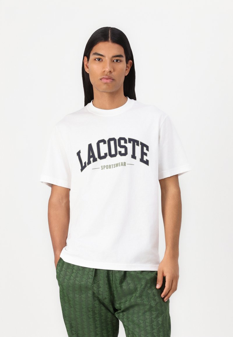 Vit bomull t-shirt med en stor marinblå "LACOSTE" logotyp och "SPORTSWEAR" i mindre text, kombinerad med gröna mönstrade byxor.