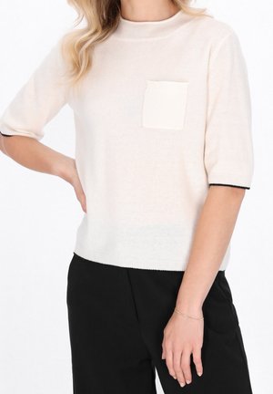 Femme portant un pull crème à manches courtes avec une poche avant et des poignets bordés de noir, associé à un pantalon noir, main sur la hanche et bracelet au poignet.
