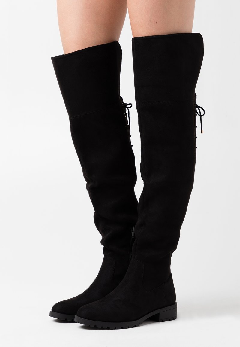 Botas de ante negras por encima de la rodilla con tacón plano, cremallera lateral y detalles de cordones en la parte trasera, con una textura suave y un diseño elegante.
