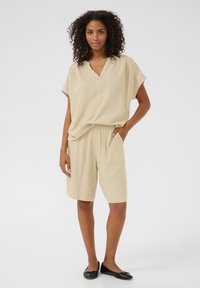 Haut beige à col en V avec des manches courtes, associé à un short assorti jusqu'aux genoux avec une taille élastique et des poches latérales. Chaussures plates noires.