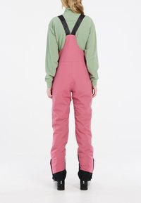 Roze waterdichte overall met verstelbare zwarte banden, voorzien van een taps toelopend ontwerp en versterkte onderbenen, gecombineerd met een groene longsleeve.