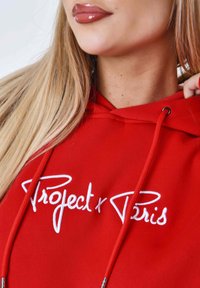 Felpa rossa con cappuccio caratterizzata da testo ricamato bianco "Project x Paris." Include cordini e una texture morbida. Dettaglio ravvicinato del collo e dei capelli.