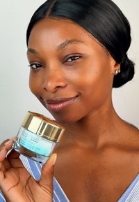 Fuktgivande kräm i en glasburk med guldlid, med en blå etikett. Personen håller den och visar upp en slät hudtextur.