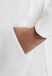 Camisa blanca con una tela texturada, que presenta un bolsillo visible y mangas remangadas, destacando un diseño suave y limpio.