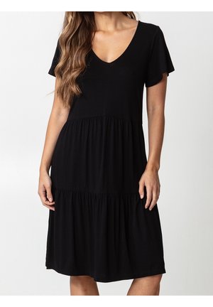 Vrouw die een zwarte jurk draagt die tot op de knie reikt, met korte mouwen, een V-hals en een gelaagde rok, staand tegen een effen achtergrond.