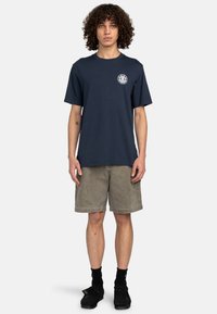 Camiseta de algodón azul marino de mangas cortas y cuello redondo; presenta un pequeño logotipo circular en el pecho. Combinada con pantalones cortos beige y zapatos negros.