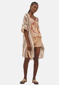 Kimono ligero y estampado en beige con acentos geométricos marrones, llevado sobre una blusa sin mangas floral y pantalones cortos beige, complementado con sandalias.