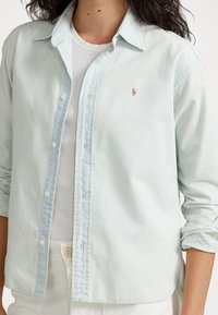 Ljusblå skjorta med knappar, gjord av mjukt tyg, med krage, långa ärmar och broderad logotyp på bröstet. Vit undertröja synlig.