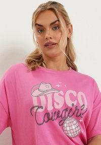 Rosa oversized t-shirt med en grafik som har texten "DISCO Cowgirl", en cowboyhatt och en discokula i en lekfull design.