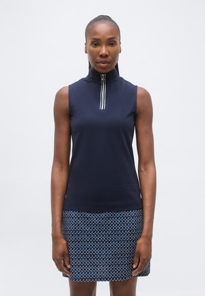 Woman stands facing forward wearing a sleeveless navy top with a front zipper and a patterned blue, black, and white skirt.