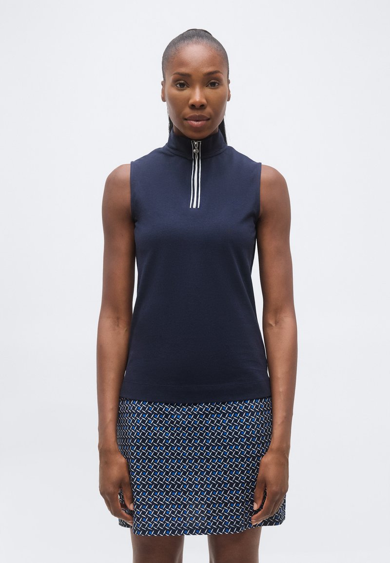 Μια γυναίκα στέκεται με το πρόσωπο προς τα εμπρός, φοράει ένα αμάνικο μπλε σκούρο τοπ με φερμουάρ μπροστά και μια πολύχρωμη φούστα με μπλε, μαύρο και λευκό μοτίβο.