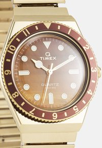 Montre à quartz Timex Q avec un boîtier en métal doré, un cadran marron, une lunette bordeaux et des index horaires blancs sur un bracelet en métal doré.