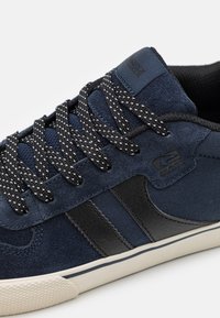 Navy suede sneakers feature black leather accents, patterned black and white laces, and a textured sole with a subtle logo.