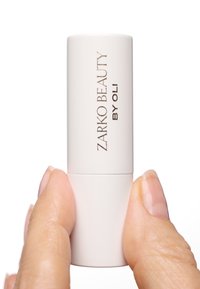 Prodotto per le labbra cilindrico bianco tenuto tra le dita, con "ZARKO BEAUTY BY OLI" in testo dorato, superficie liscia e finitura opaca.