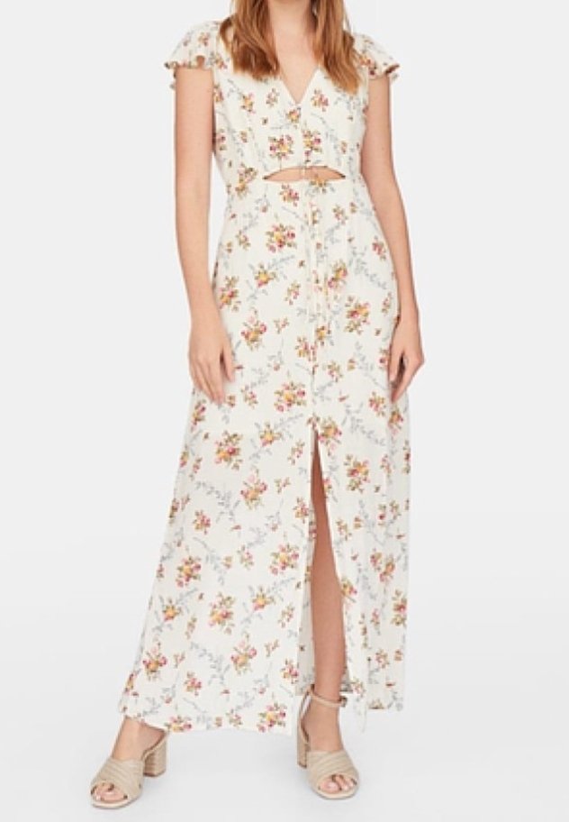Femme portant une robe maxi blanche à fleurs avec une fente à l'avant, un détail découpé à la taille et des sandales à talons beiges.