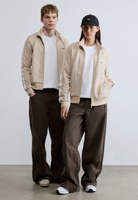 Μπεζ μπουφάν με φερμουάρ και γεωμετρικό σχέδιο, λευκές λεπτομέρειες, συνδυασμένα με λευκά T-shirt και καφέ παντελόνες φαρδιάς γραμμής, με μαύρα καπέλα και αθλητικά παπούτσια.