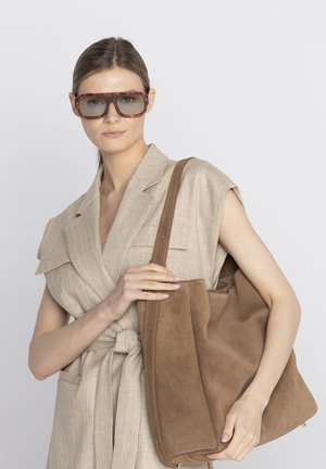 Kobieta nosząca duże brązowe okulary przeciwsłoneczne i beżową bezrękawnikową marynarkę z paskiem, trzymająca dużą brązową zamszową torbę na jednym ramieniu.