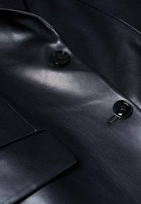 Veste en cuir noire avec texture visible, montrant deux boutons noirs et un rabat de poche avant en vue rapprochée.