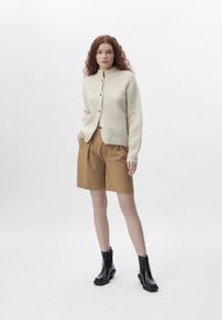 Cream knitted cardigan with buttons, paired with tailored brown shorts and black ankle boots. Smooth fabric texture and relaxed fit.