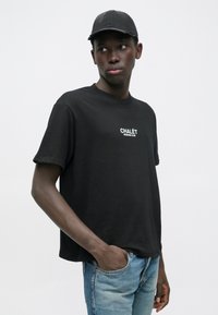 Černé bavlněné tričko s krátkými rukávy a kulatým výstřihem, s bílým nápisem "CHALÉT SPORTING CLUB". Kombinujte s světle modrými džínami.