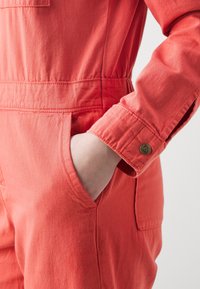 Combinaison en coton rouge avec une taille cintrée, des manches longues et une poche latérale. Elle est dotée d'un bouton métallique de marque sur le poignet.