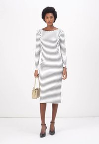 Robe midi rayée en noir et blanc, à manches longues, décolleté bateau classique, silhouette ajustée, portée avec des talons noirs et un sac à main beige.