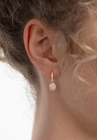 Boucles d'oreilles créoles en or avec un pendentif ovale orné de petites pierres scintillantes. Vue en gros plan mettant en valeur la texture et l'éclat.