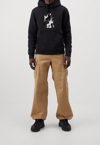 Felpa con cappuccio nera con stampa grafica e logo, abbinata a pantaloni cargo kaki con tasche laterali e scarpe sportive nere.