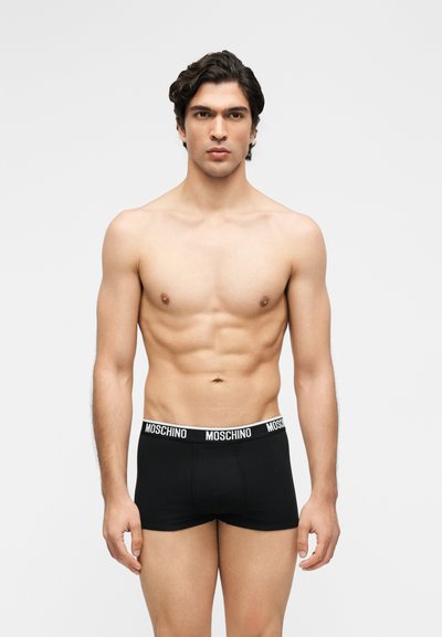 Νέος άνδρας με σκούρα μαλλιά που φοράει μαύρα μποξεράκια Moschino, στέκεται μπροστά σε απλό φωτεινό φόντο.