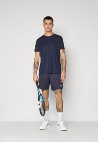 Marinblå träningströja med korta ärmar, i kombination med mörka shorts. Motivet håller en tennisklubba, iförd vita strumpor och sneakers.