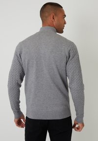 Šed�ý pletený svetr s vysokým límcem, s texturovanými vzorovanými rukávy. Tkanina má hladký vzhled, což kontrastuje s detailními akcenty.