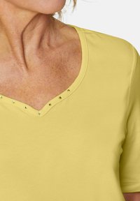 Gelbes V-Ausschnitt-T-Shirt mit kurzen Ärmeln, verziert mit metallischen Nieten am Kragen. Weicher Stoff mit glatter Textur.