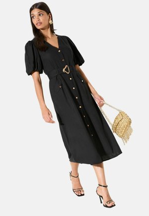 Nő fekete midi ruhában, amely puffos rövid ujjú, elöl gombos, széles övvel, fekete magassarkú szandálban, és egy arany szövött táskát tart a kezében.