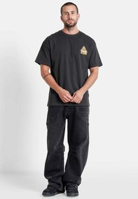 Zwarte katoenen t-shirt met een goudkleurige grafiek op de borst, gecombineerd met losse zwarte denimbroeken en zwart schoeisel.