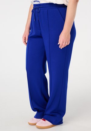 Blauwe, ruime sweatpants met een elastische tailleband en een trekkoord. Voorzien van zijzakken en een gestructureerde stof. Gecombineerd met roze sneakers.