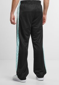 Crne trenirke s svijetloplavim bočnim panelom koji ima geometrijski uzorak. Elastičan pojas i gladak tekstur. Bijele tenisice su vidljive.