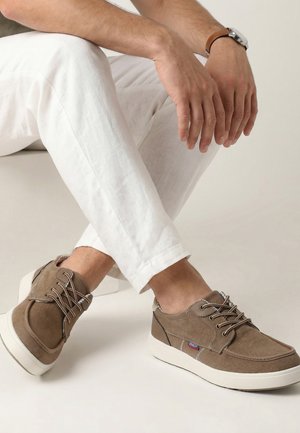 Mann trägt beige Schnürschuhe und weiße Hose, sitzt mit überkreuzten Beinen und legt die Hände auf das Knie, vor weißem Hintergrund.